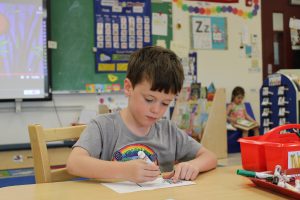 pre-K student draws with markers. Another student reads a book in the background