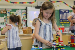 pre-K girl builds with legos while other students play behind her as well