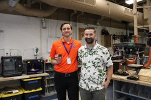 Evan Lally and Charles Davis smile for the camera. Behind them are 3D printers and traditional equiptment. Lally holds a micro:bit