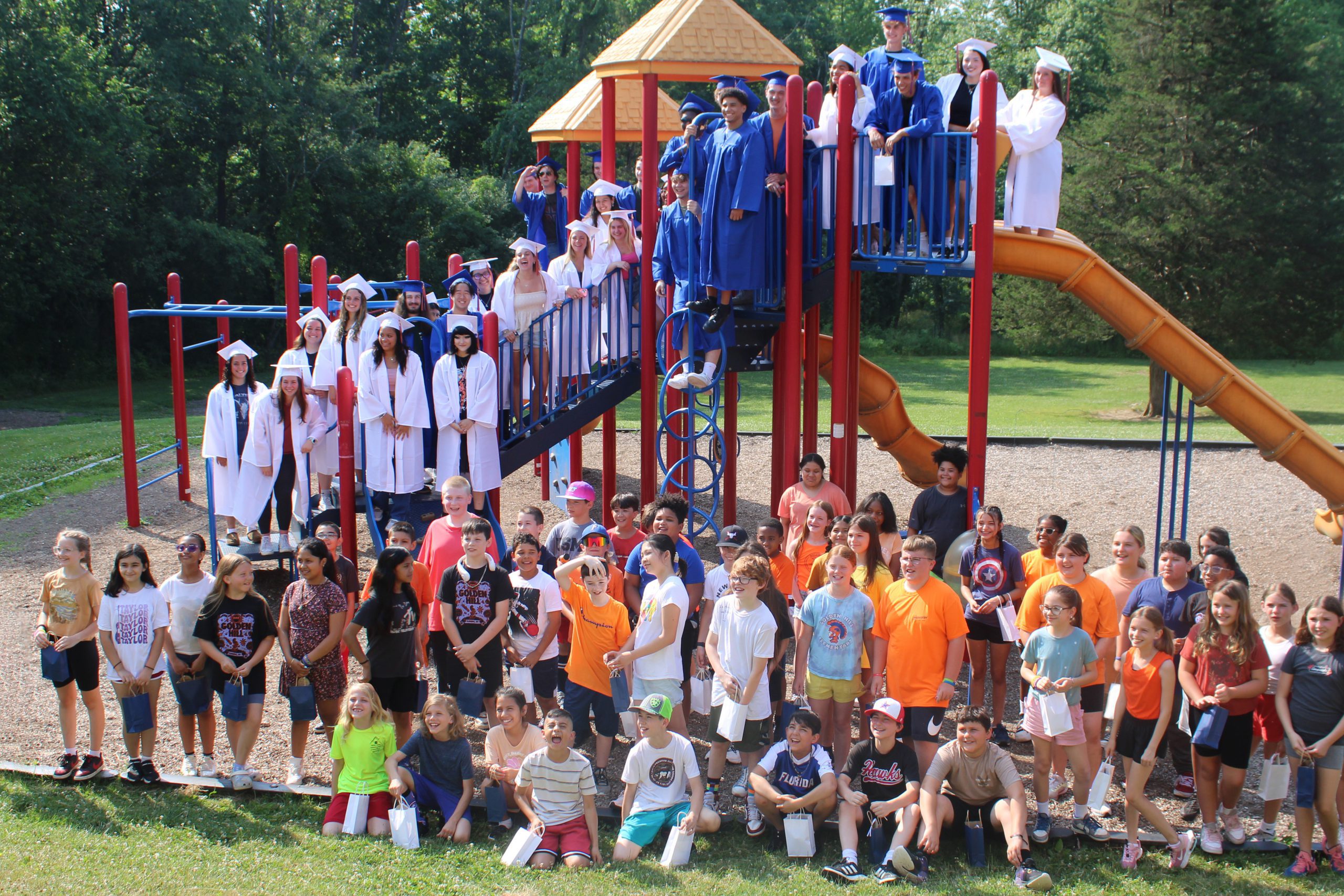 Seniors, in caps and gown, stand on the playground smiling with 5th graders down below