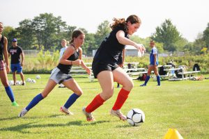 A girl dribbles a soccer ball, closely followed by a challenging player