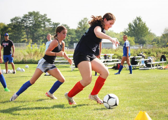 A girl dribbles a soccer ball, closely followed by a challenging player