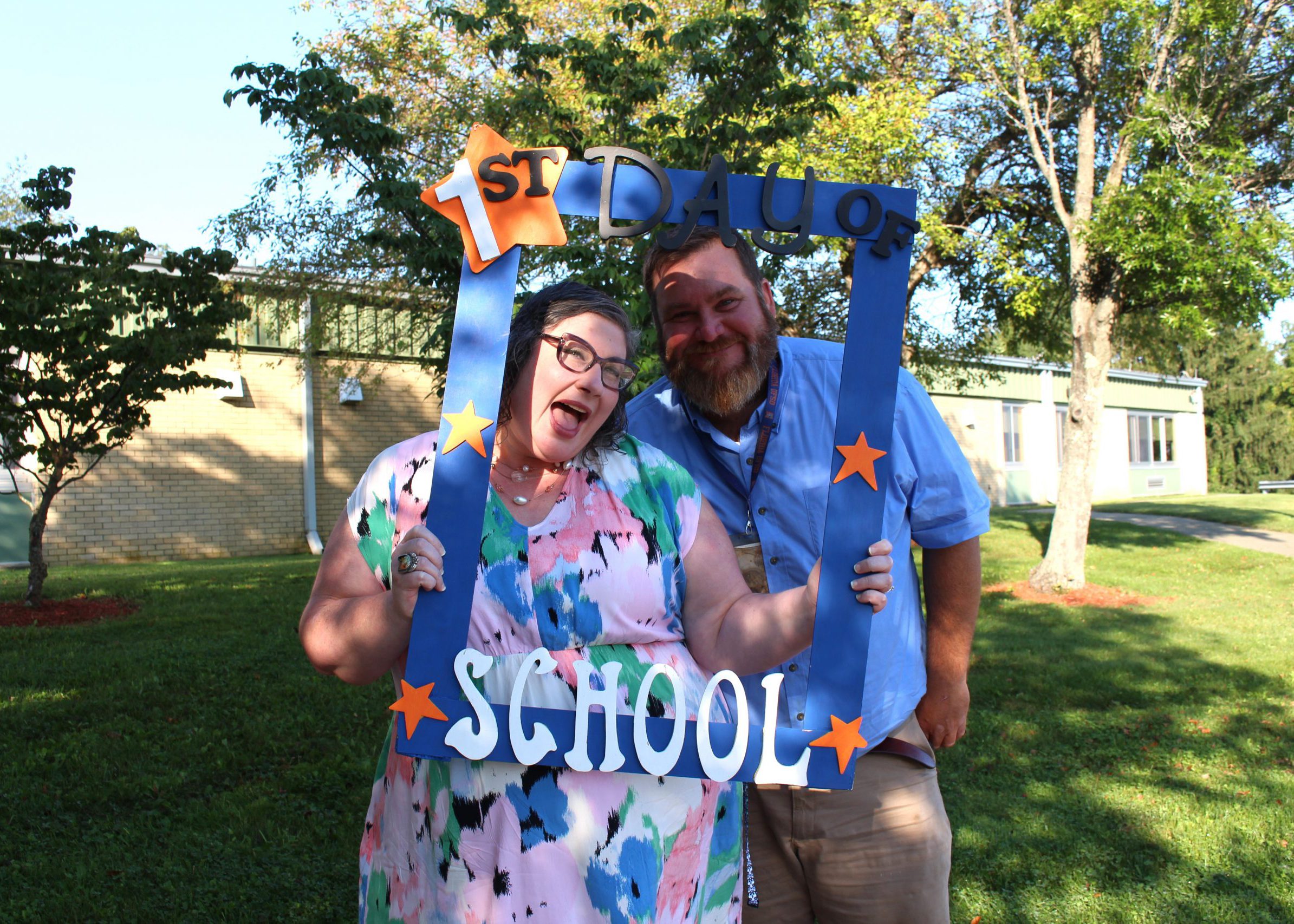 two staff members smile from inside large frame that says "first day of school"