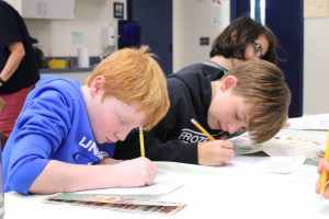 two students sit at table covered with tablecloth for decimal restaurant event, working with local menus to complete word problems